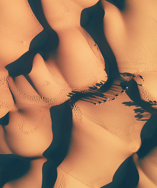 Aerial view of sandy dunes in the desert with lines of people riding camels.